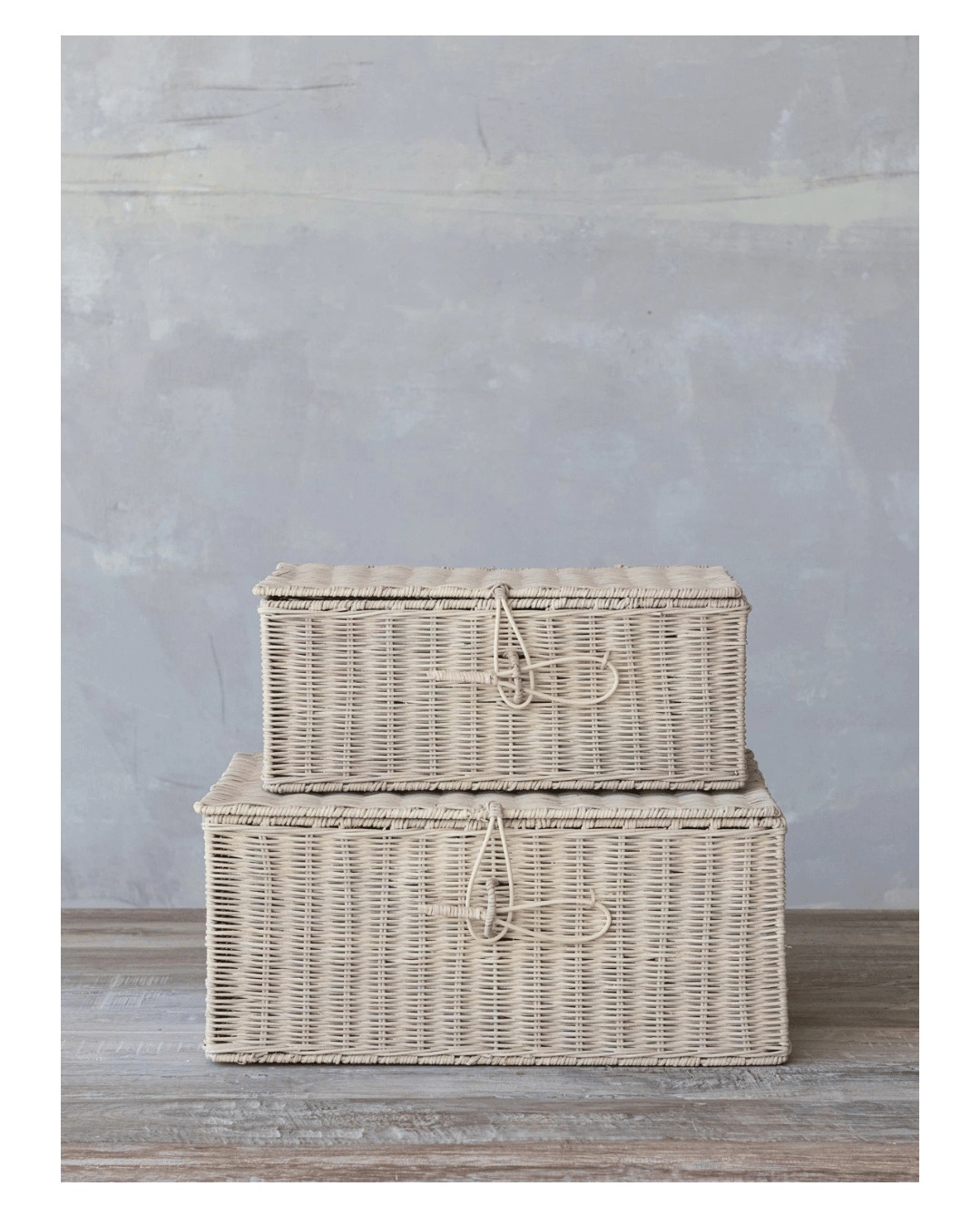 Two hand-woven rectangular rattan boxes with closures from Creative Co-op, bleached and featuring simple loop closures, are stacked on a wooden floor against a textured gray wall.