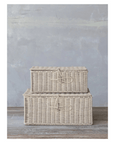 Two hand-woven rectangular rattan boxes with closures from Creative Co-op, bleached and featuring simple loop closures, are stacked on a wooden floor against a textured gray wall.