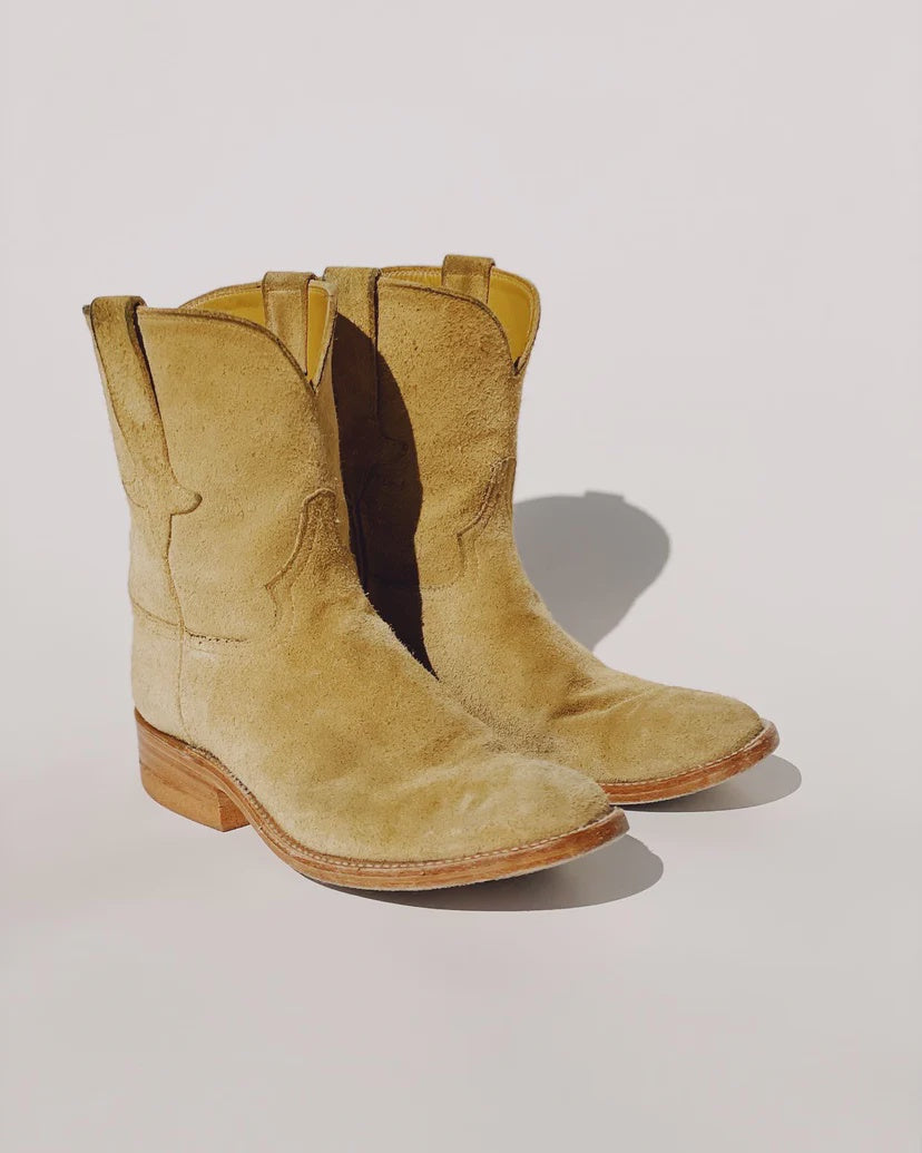 A pair of "The Ranch Boot" by Hey Gang in light brown calfskin suede, featuring leather soles, is positioned side by side on a plain white background. The soft texture and classic western style are clearly visible.