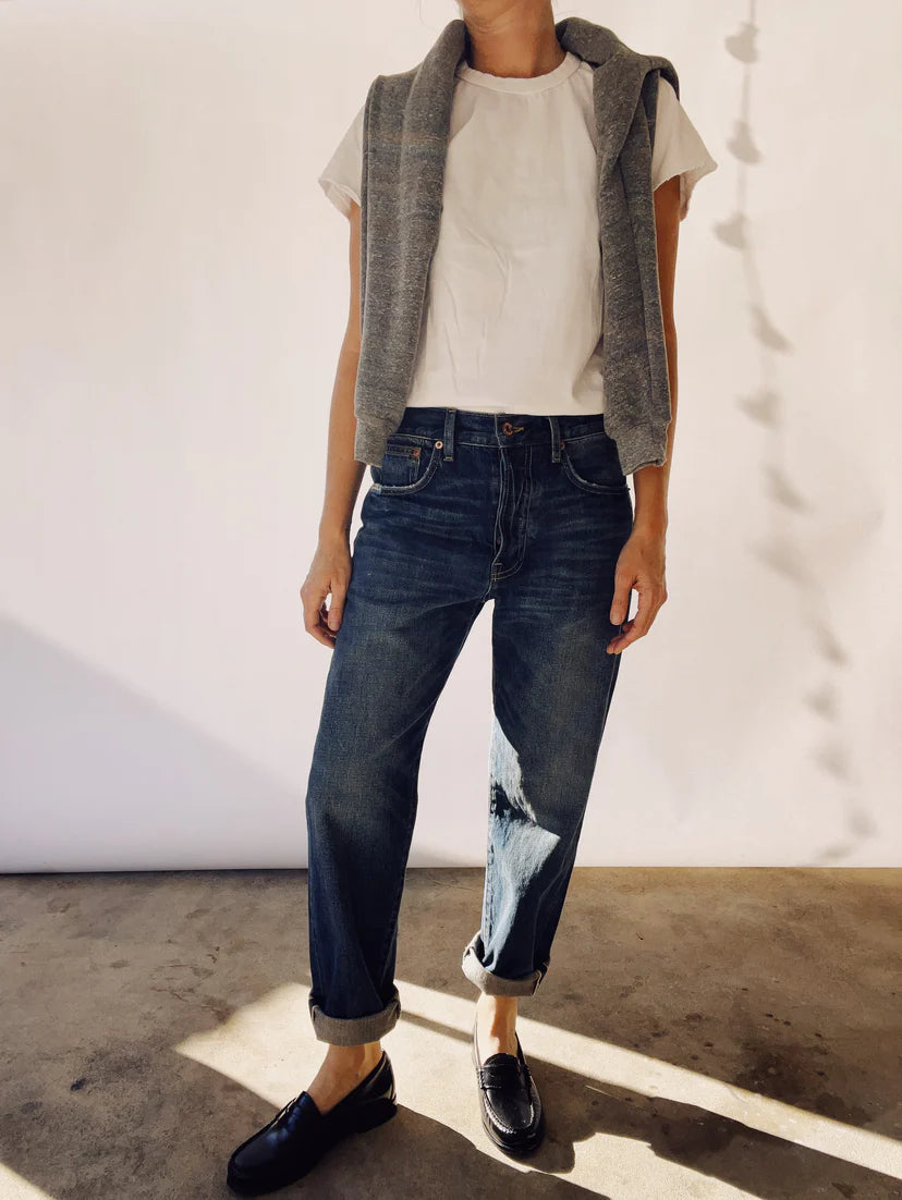 A person stands casually wearing cuffed "The Loose Crop Selvedge" by Hey Gang, paired with a white T-shirt and a grey sweater draped over their shoulders. They are also wearing black loafers against a plain white wall with shadows.