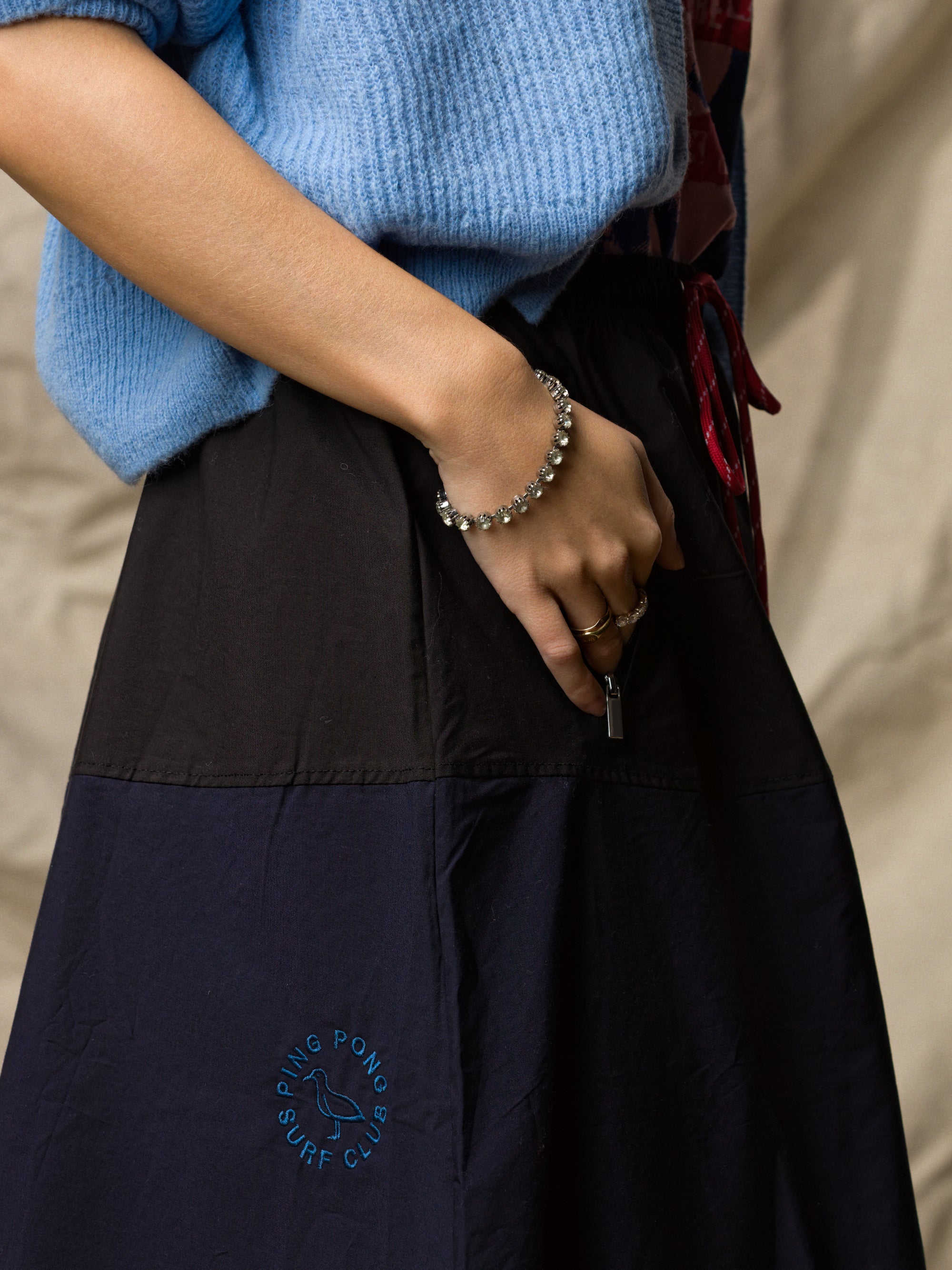 A person wears the Le Vintage Sport Skirt by Ping Pong Surf Club and a blue sweater, resting one hand in their pocket to reveal a pearl bracelet and rings. The backdrop is a neutral, textured fabric.