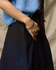 A person wears the Le Vintage Sport Skirt by Ping Pong Surf Club and a blue sweater, resting one hand in their pocket to reveal a pearl bracelet and rings. The backdrop is a neutral, textured fabric.