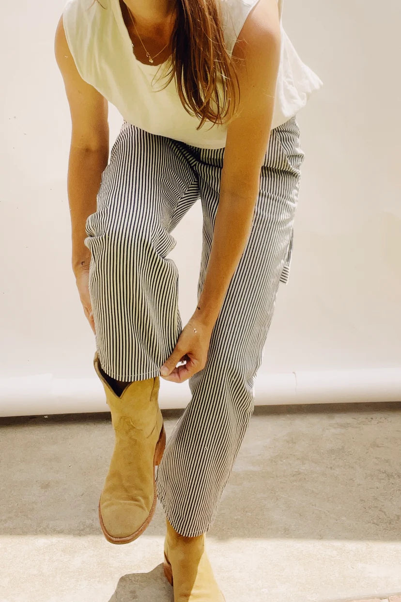 A person wearing a white sleeveless top and striped pants is adjusting their Hey Gang's The Ranch Boot, made of tan calfskin suede in a roper style. They are bent over slightly, situated in a well-lit area with a neutral backdrop that creates a casual and relaxed atmosphere.