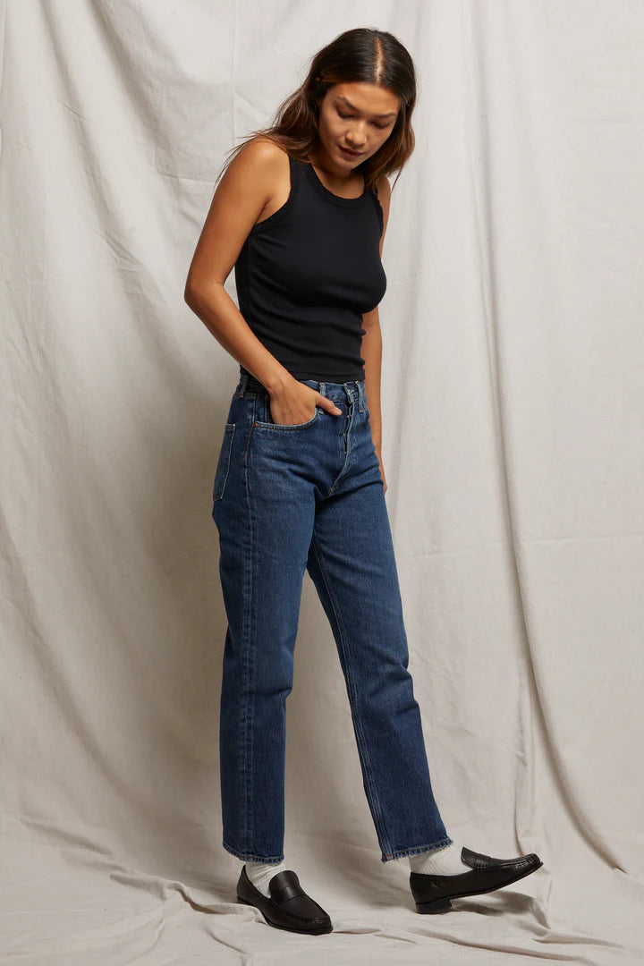 A person stands against a light fabric backdrop wearing the Elle Rib Tank in black by Perfectwhitetee, paired with blue high-waisted jeans, white socks, and black loafers. One hand is in their pocket while the other hangs by their side as they glance down slightly.