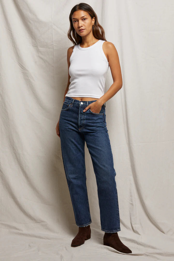 Against a light backdrop, a person showcases their long, wavy hair while wearing the bra-friendly Elle Rib Tank by Perfectwhitetee. This versatile white sleeveless top is paired with blue jeans and brown boots, with one hand resting in their pocket.