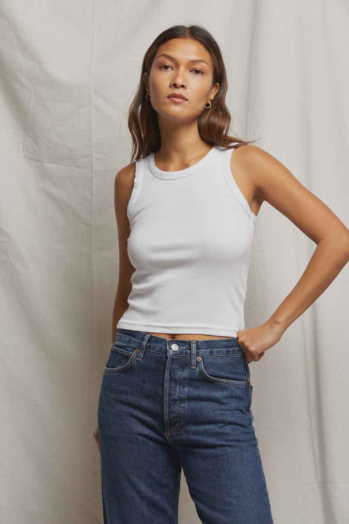 A person with long hair confidently poses against a plain backdrop, wearing the versatile Elle Rib Tank from Perfectwhitetee and blue jeans, with their left hand on their hip.