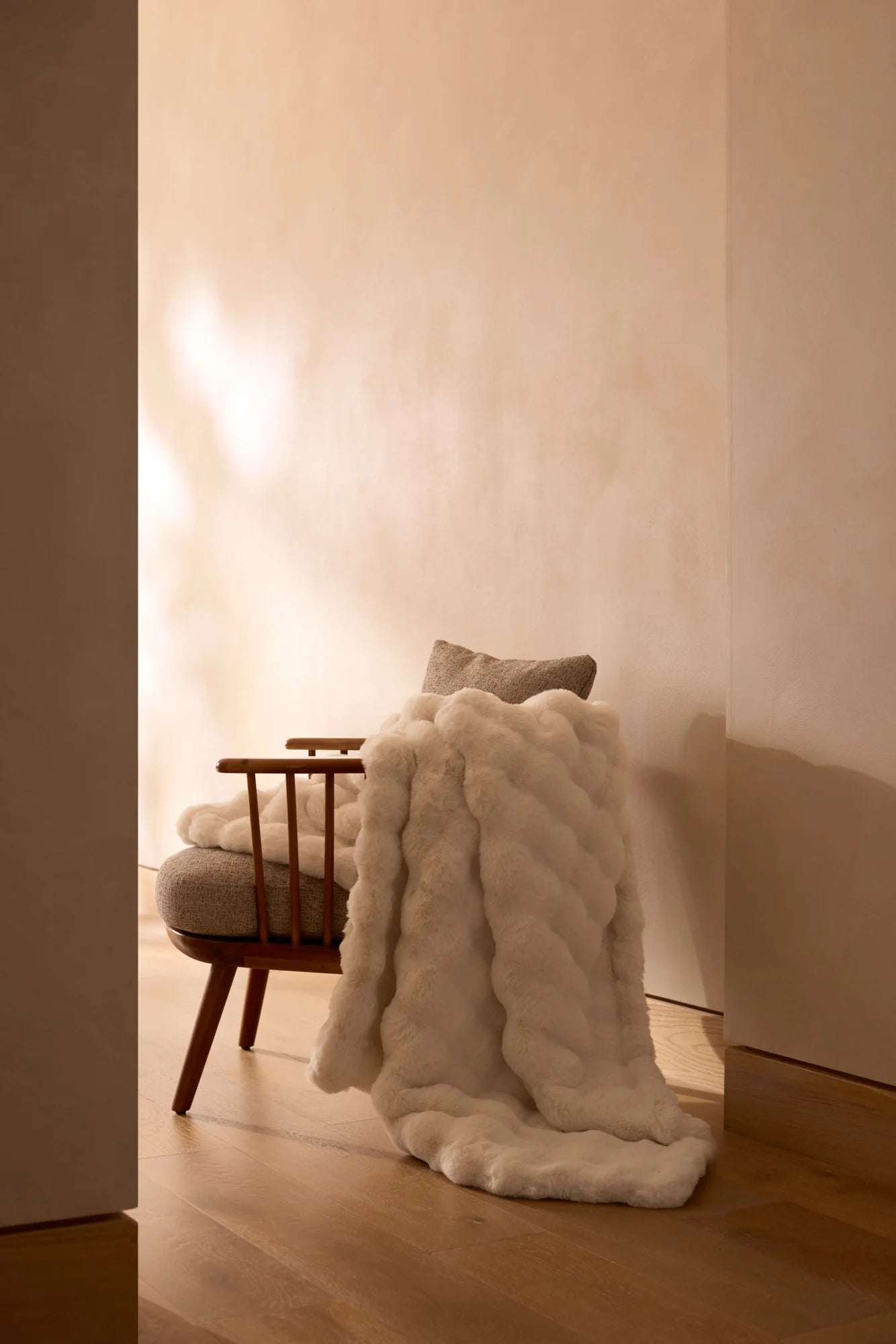 A Cozy Earth Bubble Cuddle Blanket, crafted from ultra-soft fibers, is draped over a cushioned wooden chair with a neutral pillow, set in a minimal, warmly lit room with a beige wall and wooden floor.