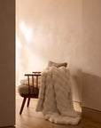 A Cozy Earth Bubble Cuddle Blanket, crafted from ultra-soft fibers, is draped over a cushioned wooden chair with a neutral pillow, set in a minimal, warmly lit room with a beige wall and wooden floor.