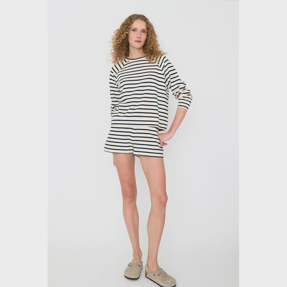 A person with long curly hair stands against a plain background, wearing Kule's Eco Cotton Venus set, featuring a striped long-sleeve shirt and matching pull-on shorts made of organic cotton. Their neutral expression is complemented by beige slip-on shoes.