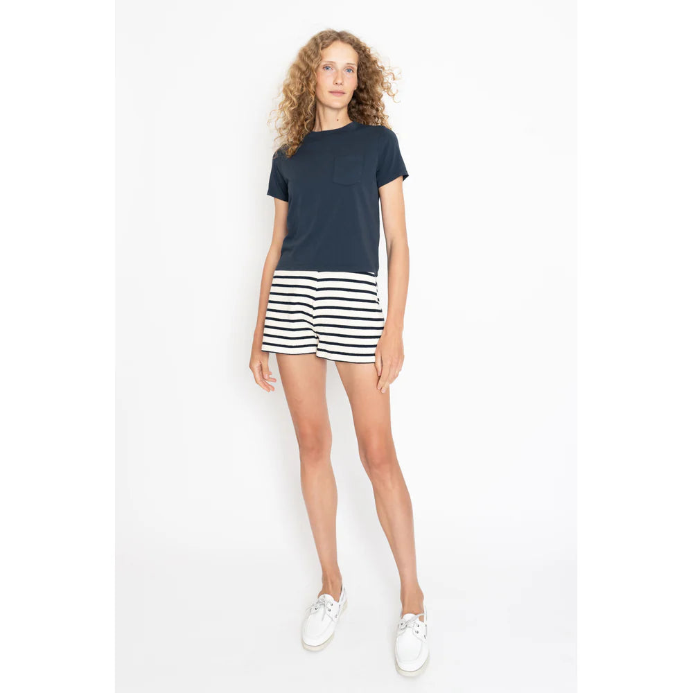 A curly-haired person wearing a navy t-shirt, The Eco Cotton Venus by Kule as black and white striped pull-on shorts, and white sneakers stands against a plain white background.