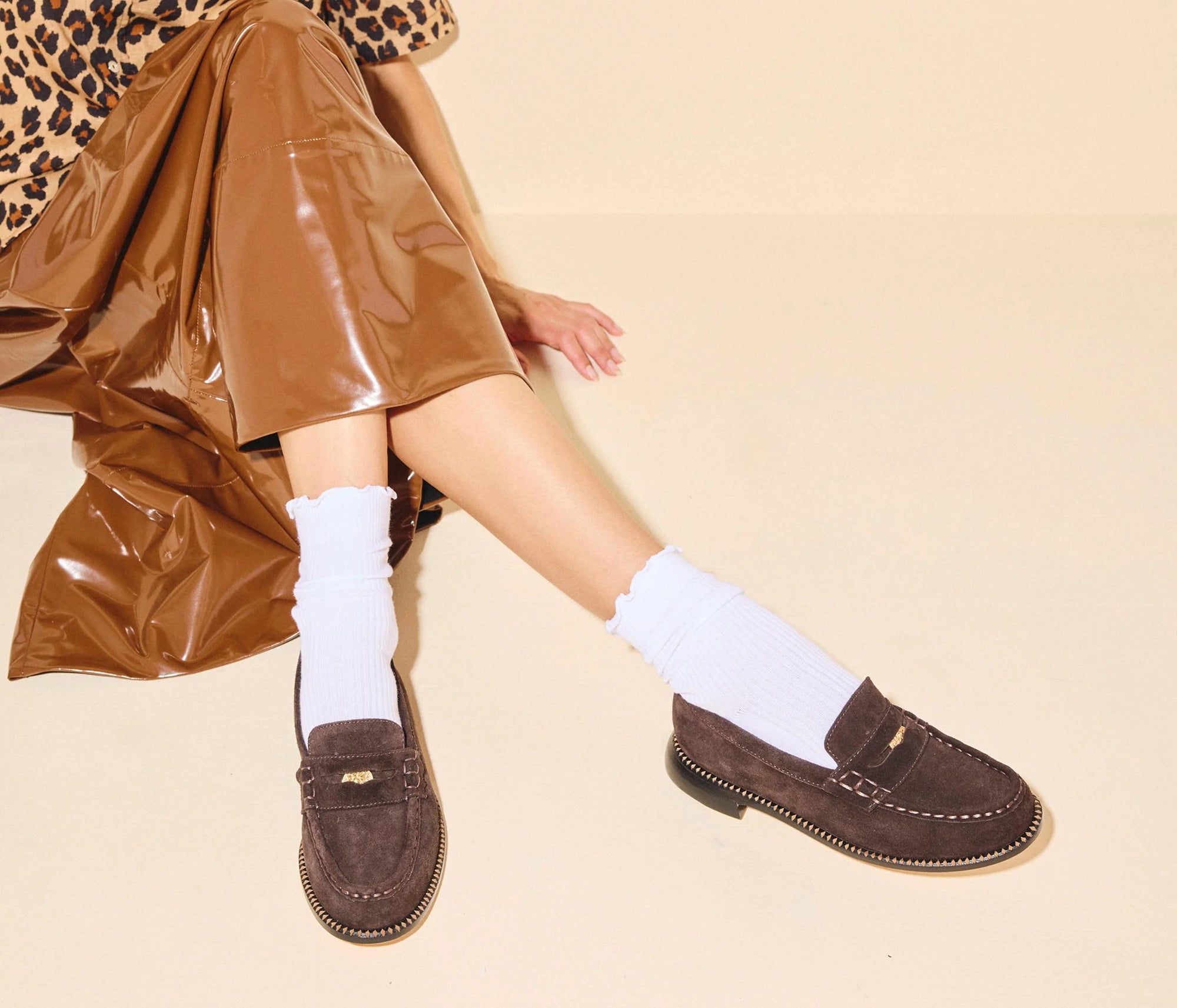A person sits cross-legged on a cream background in a brown leopard-print top, shiny brown skirt, white ribbed socks, and Freda Salvador's Elba Espresso Suede Loafer.
