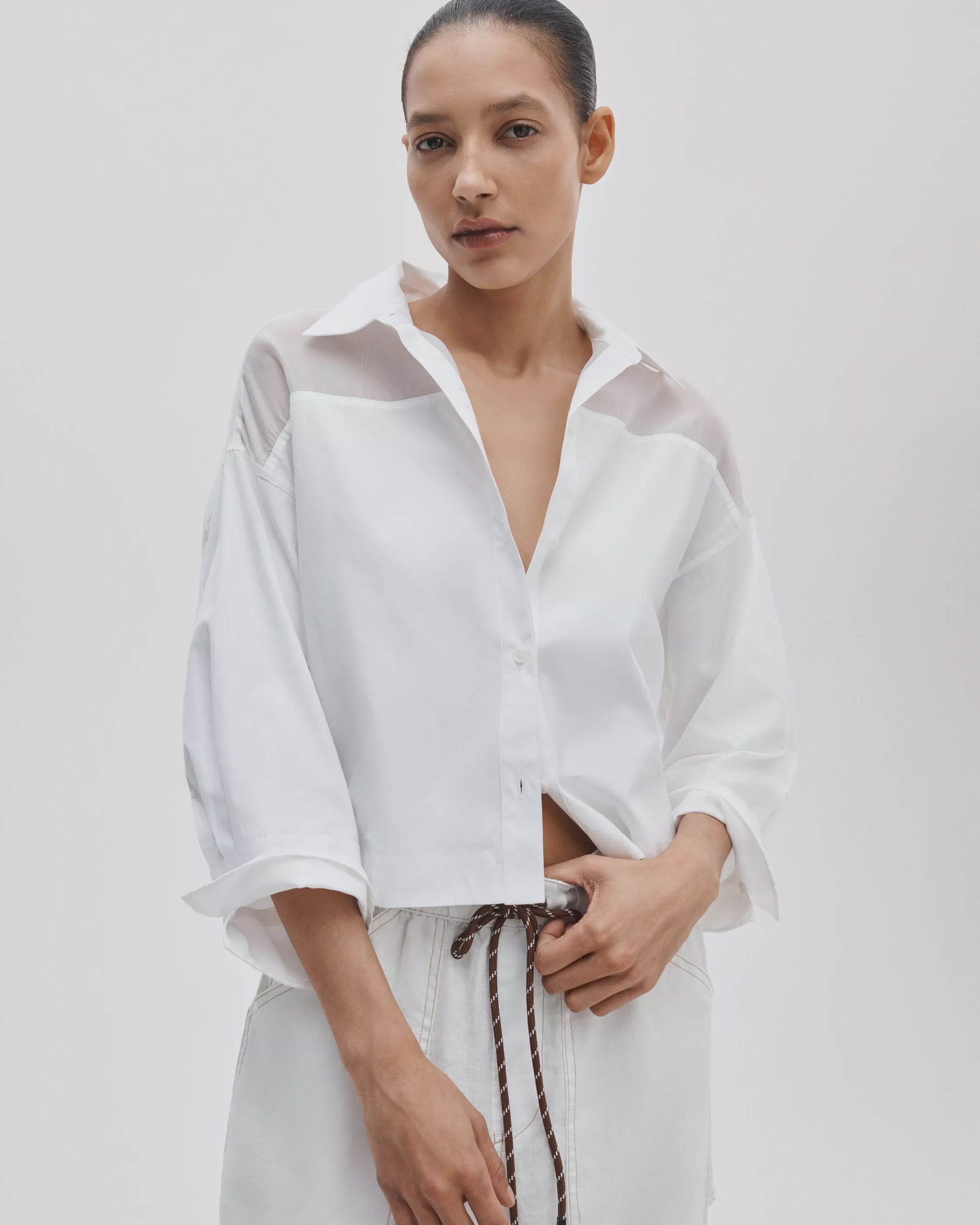 A woman wears the Rag & Bone elly ss shirt—a cropped, boxy cotton poplin top with semi-sheer organza shoulder panels—paired with white pants and a brown rope belt, standing against a plain light background.