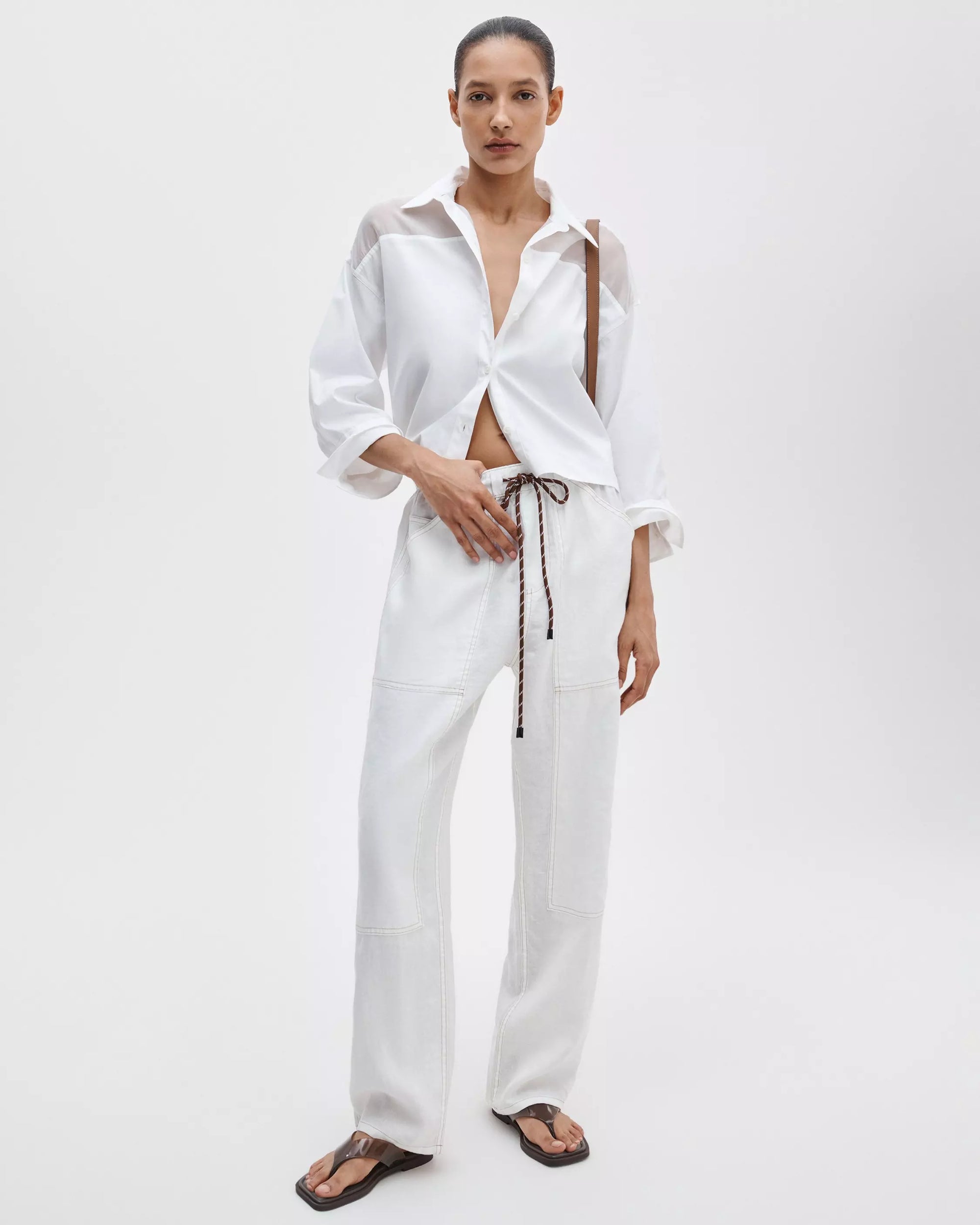 A woman poses with one hand in her pocket against a white background, wearing the Rag & Bone elly ss shirt—a cropped, boxy, semi-sheer organza top—paired with loose drawstring pants, flip-flop sandals, and a crossbody bag.