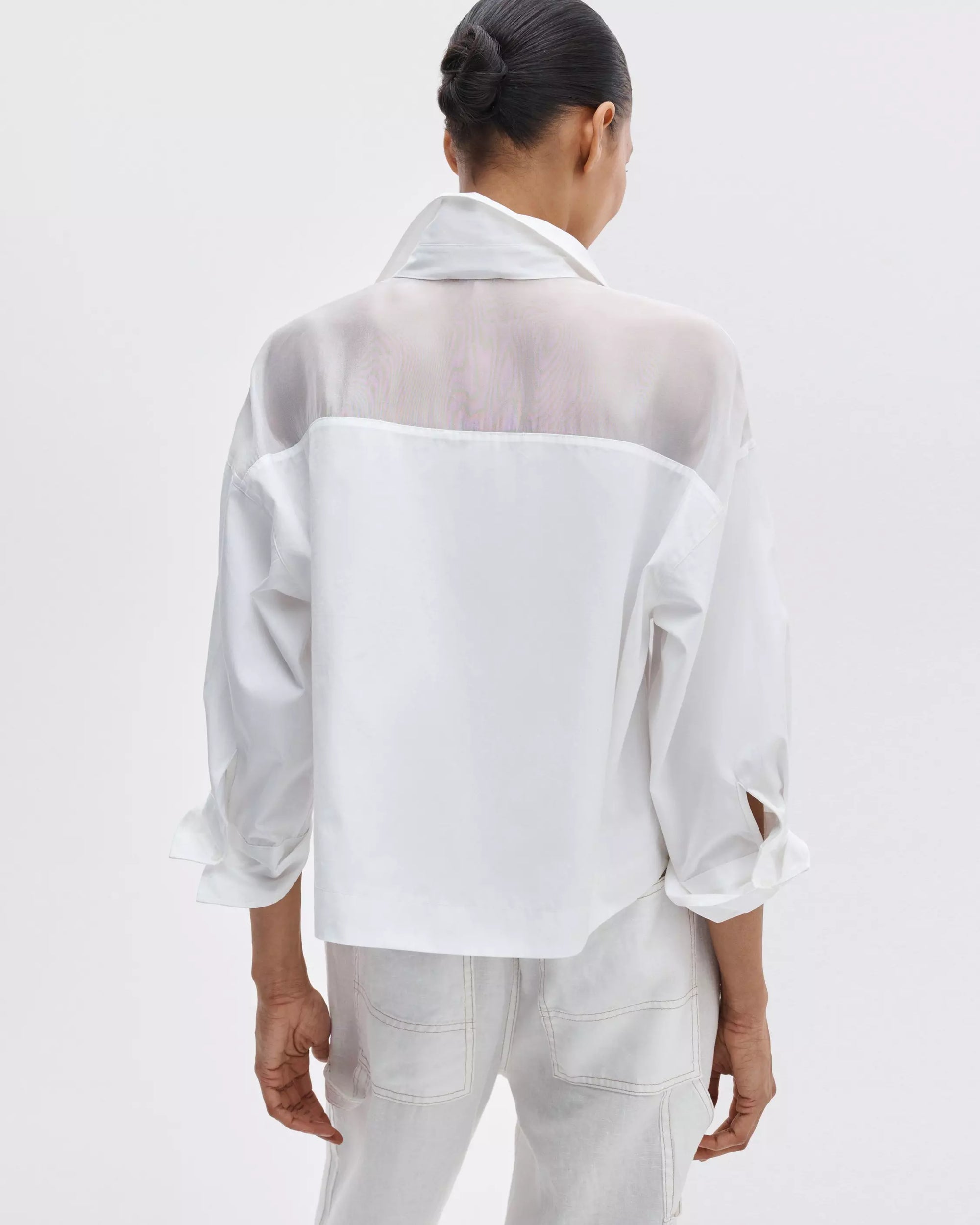 A woman with dark hair in a bun faces away from the camera, wearing the Rag & Bone elly ss shirt—a semi-sheer white organza blouse with rolled sleeves—paired with white jeans against a plain light background.