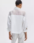 A woman with dark hair in a bun faces away from the camera, wearing the Rag & Bone elly ss shirt—a semi-sheer white organza blouse with rolled sleeves—paired with white jeans against a plain light background.