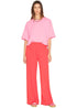 A person with straight brown hair and a neutral expression stands against a white background, wearing a loose pink T-shirt, Xirena Emmette Sweatpant in bright red, and pink shoes.