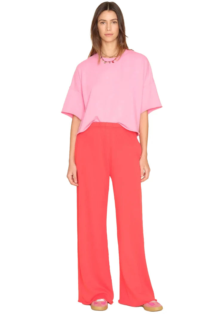 A person with straight brown hair and a neutral expression stands against a white background, wearing a loose pink T-shirt, Xirena Emmette Sweatpant in bright red, and pink shoes.
