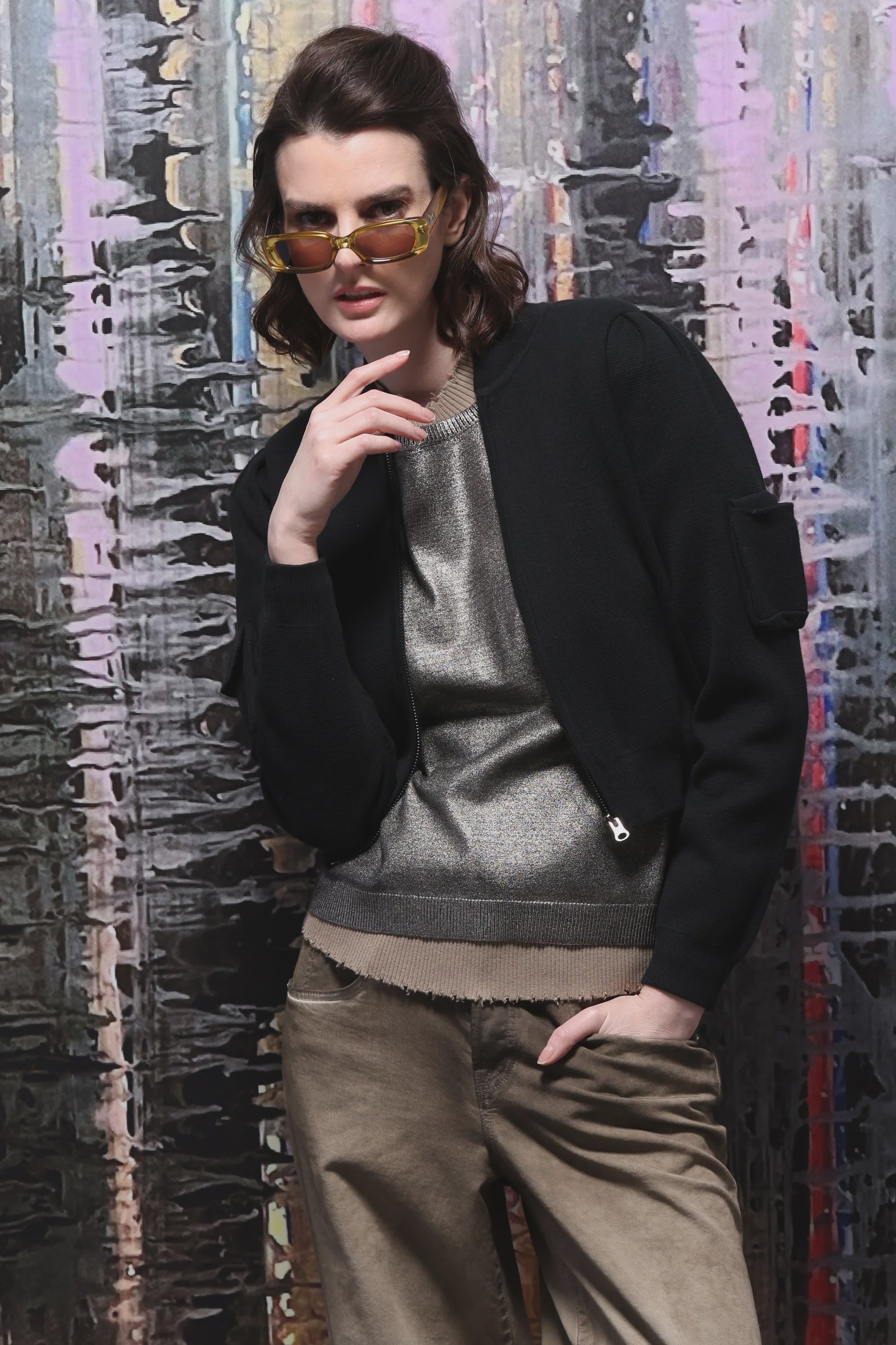 A person wearing sunglasses, a Cropped Bomber Jacket by Minnie Rose, and a luxurious cotton blend metallic top paired with khaki pants poses with one hand on their chin. The background features a colorful abstract pattern with vertical brushstrokes.