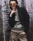 A person wearing sunglasses, a Cropped Bomber Jacket by Minnie Rose, and a luxurious cotton blend metallic top paired with khaki pants poses with one hand on their chin. The background features a colorful abstract pattern with vertical brushstrokes.