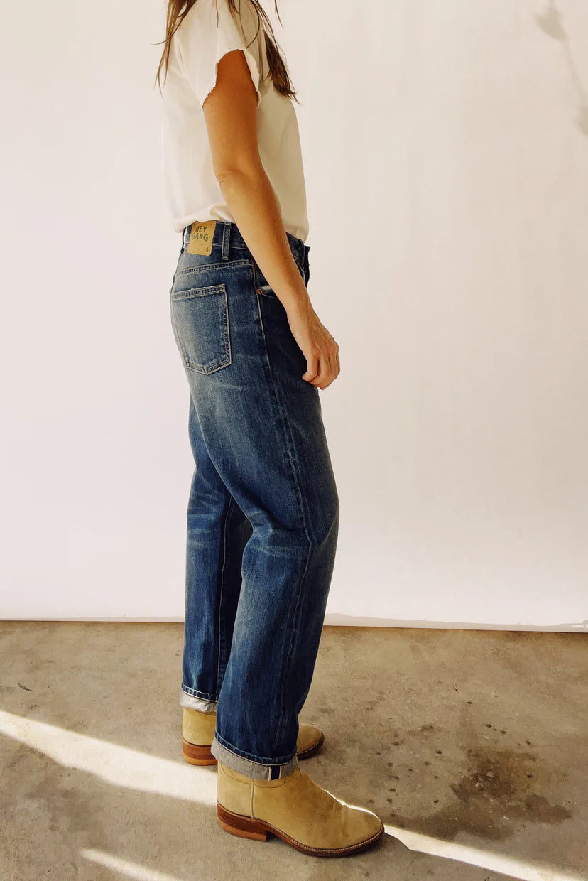 A person stands against a white background, dressed in The Loose Crop Selvedge jeans by Hey Gang and a white shirt, paired with beige boots. They face slightly away from the camera, casting subtle light and shadows on the concrete floor.