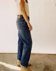 A person stands against a white background, dressed in The Loose Crop Selvedge jeans by Hey Gang and a white shirt, paired with beige boots. They face slightly away from the camera, casting subtle light and shadows on the concrete floor.