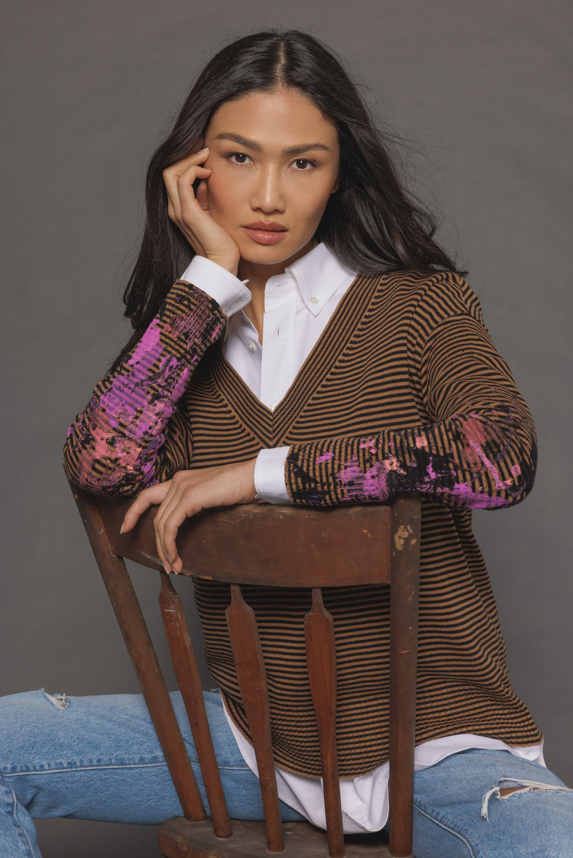 Seated on a wooden chair with long dark hair facing backward, the person is dressed in Lisa Todd's "Between the Lines" sweater layered over a white shirt and paired with ripped jeans against a plain gray backdrop.