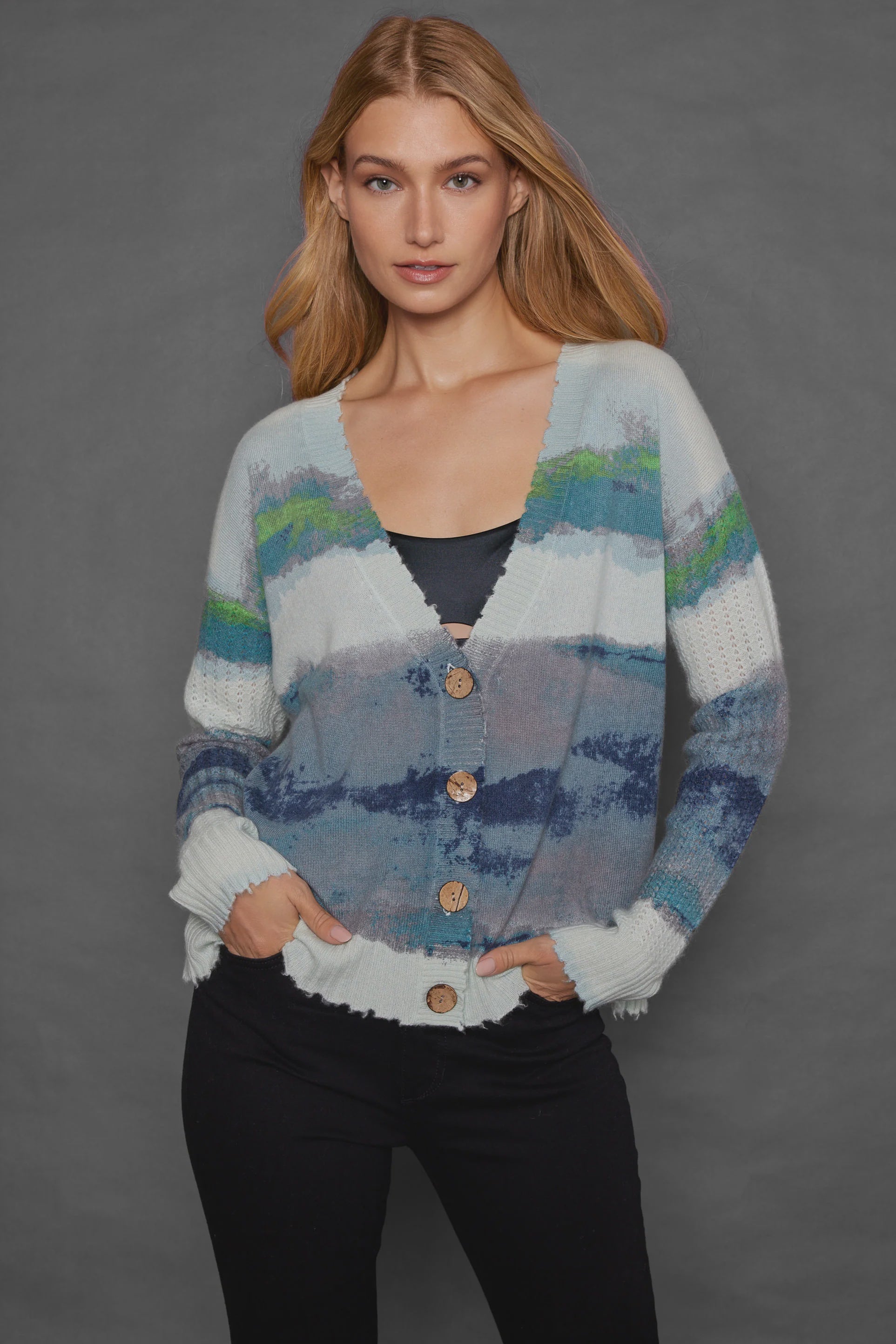 A woman with long blonde hair is wearing Lisa Todd's Color Coded Cardigan, which showcases a brush stroke print in blue, green, and gray stripes with front buttons. She has her hands tucked into the pockets of relaxed fit black pants while standing against a plain gray background.