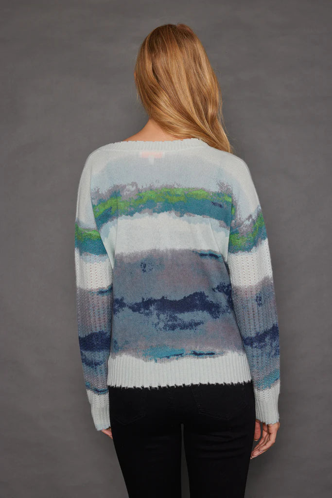 A person with long blonde hair stands facing away, wearing the Lisa Todd Color Coded Cardigan featuring horizontal brush stroke print in shades of blue, green, and gray. They are paired with black pants against a plain, dark background.