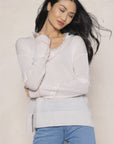 A person with long hair stands confidently, wearing Lisa Todd's "In Fringe," a light-colored V-neck pullover featuring subtle fringe details, paired with blue jeans. They tilt their head slightly with a gentle smile, embodying a casual and relaxed vibe against a neutral background.