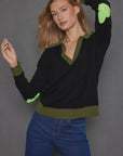 A person models the Polo Pop by Lisa Todd, a relaxed-fit black cashmere polo sweater featuring distinctive green accents on the collar, cuffs, and heart intarsia elbow patches. They pose with one hand in their hair, effortlessly pairing it with blue jeans against a gray backdrop.