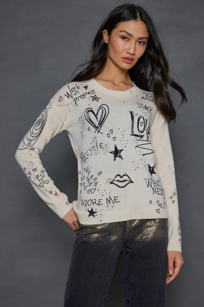 A woman poses confidently in *The Messenger Crewneck* by Lisa Todd, a gray cashmere sweater featuring black doodles of hearts, stars, and affirmations such as "Adore Me" and "Work in Progress." She complements it with metallic silver pants against a dark background.