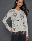 A woman poses confidently in *The Messenger Crewneck* by Lisa Todd, a gray cashmere sweater featuring black doodles of hearts, stars, and affirmations such as "Adore Me" and "Work in Progress." She complements it with metallic silver pants against a dark background.