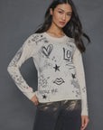 A person with long dark hair stands against a gray background wearing The Messenger Crewneck by Lisa Todd, which features doodles like hearts, stars, and uplifting messages such as "Adore Me" and "Recycle." They're also dressed in dark pants and have a relaxed pose.