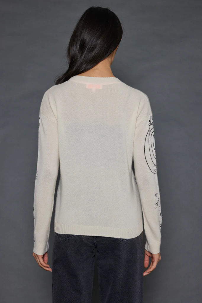 A person with long dark hair stands facing away, wearing The Messenger Crewneck by Lisa Todd, which is a light-colored cashmere sweater adorned with design details on the sleeves, paired with dark jeans against a plain, dark background.