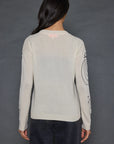 A person with long dark hair stands facing away, wearing The Messenger Crewneck by Lisa Todd, which is a light-colored cashmere sweater adorned with design details on the sleeves, paired with dark jeans against a plain, dark background.