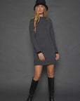 A woman with long blonde hair wearing the Lisa Todd Hide & Seek Dress, a dark grey zip neck sweater dress with contrast cuffs, stands against a grey background. She completes her look with black boots and a black hat.