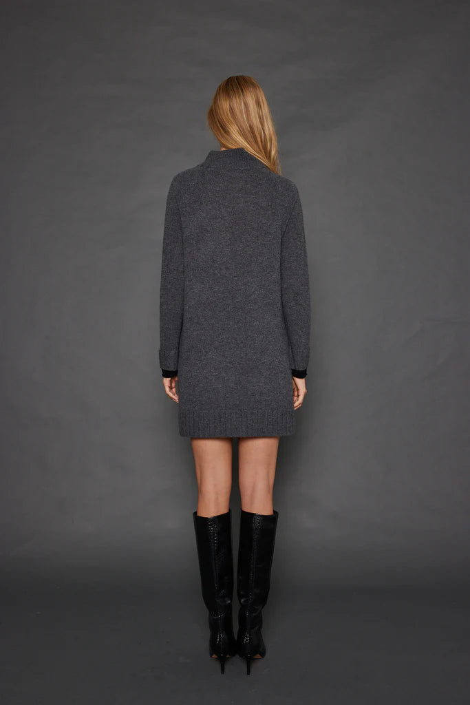 A woman with long blonde hair stands against a gray background, facing away from the camera. She is wearing the chic Hide & Seek Dress by Lisa Todd, a dark gray cashmere blend turtleneck sweater dress with contrast cuff details, paired with sleek black knee-high boots.