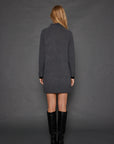 A woman with long blonde hair stands against a gray background, facing away from the camera. She is wearing the chic Hide & Seek Dress by Lisa Todd, a dark gray cashmere blend turtleneck sweater dress with contrast cuff details, paired with sleek black knee-high boots.