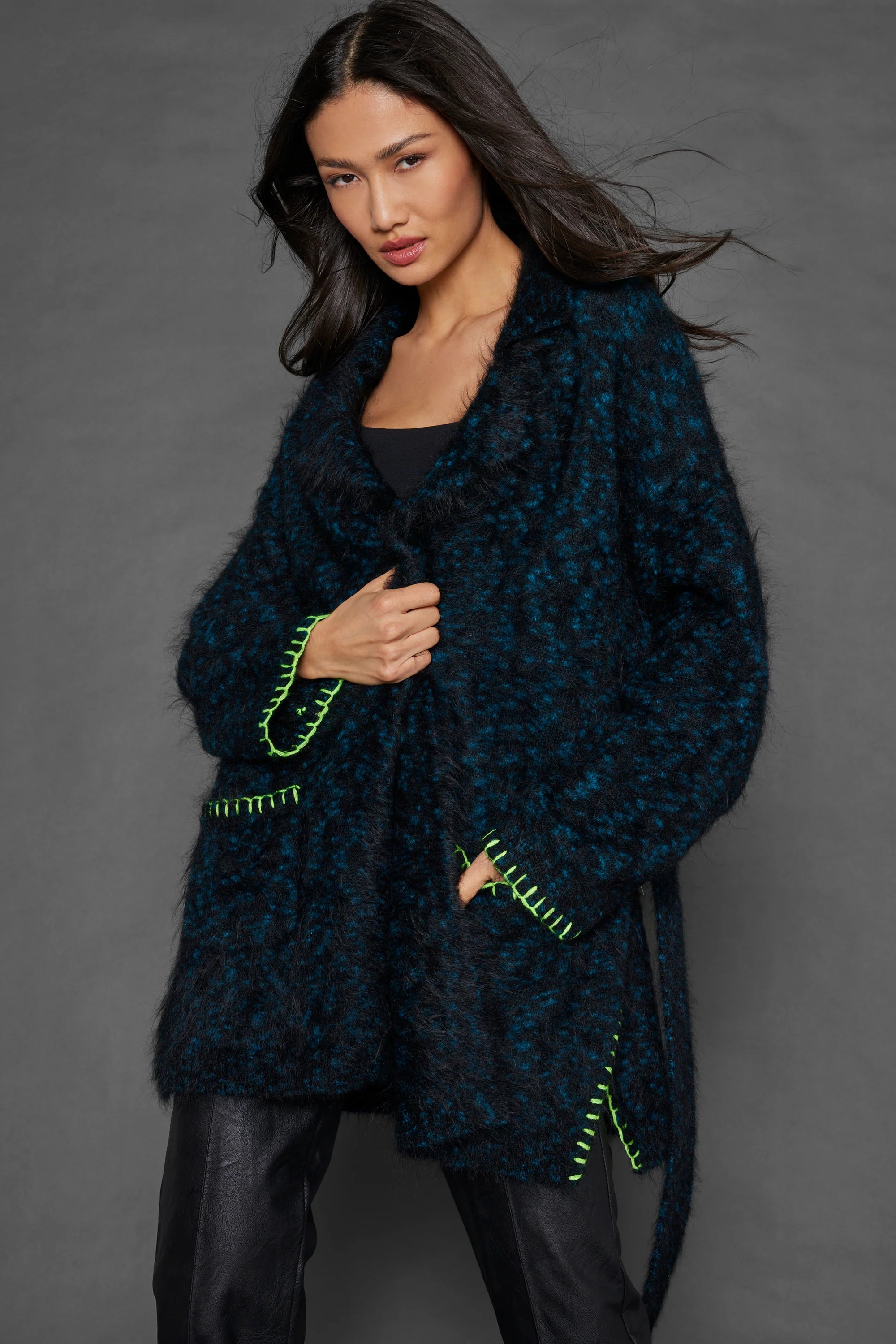 A woman with long dark hair wears the Lisa Todd Mohair Limited Edition cardigan, a blue and black textured piece crafted from Italian yarn, adorned with bright green stitched accents, layered over a black top and dark pants. She stands against a gray background with a confident expression.