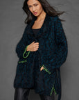 A woman with long dark hair wears the Lisa Todd Mohair Limited Edition cardigan, a blue and black textured piece crafted from Italian yarn, adorned with bright green stitched accents, layered over a black top and dark pants. She stands against a gray background with a confident expression.