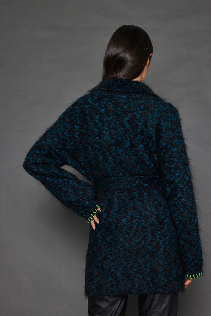 A person with long dark hair stands facing away, wearing the Mohair Limited Edition wrap knit coat by Lisa Todd. The coat features a fuzzy dark blue and black texture with a belt, subtly adorned with an animal print pattern. They have one hand on their hip while the other arm rests relaxed against a solid, neutral background.