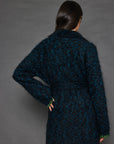 A person with long dark hair stands facing away, wearing the Mohair Limited Edition wrap knit coat by Lisa Todd. The coat features a fuzzy dark blue and black texture with a belt, subtly adorned with an animal print pattern. They have one hand on their hip while the other arm rests relaxed against a solid, neutral background.