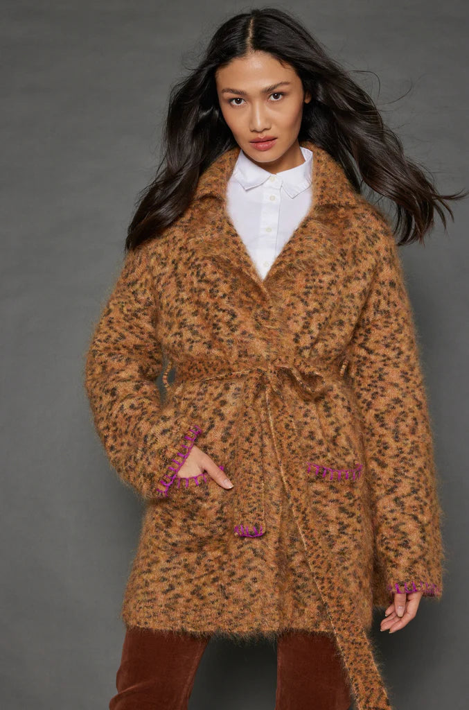 A woman with long dark hair models the "Mohair Limited Edition" by Lisa Todd—a brown fuzzy coat crafted from exquisite Italian yarn. She pairs it with a white shirt and brown pants, showcasing the coat's unique animal print complemented by pink accents along the edges, all set against a plain dark background.