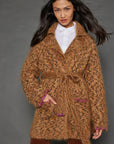 A woman with long dark hair models the "Mohair Limited Edition" by Lisa Todd—a brown fuzzy coat crafted from exquisite Italian yarn. She pairs it with a white shirt and brown pants, showcasing the coat's unique animal print complemented by pink accents along the edges, all set against a plain dark background.