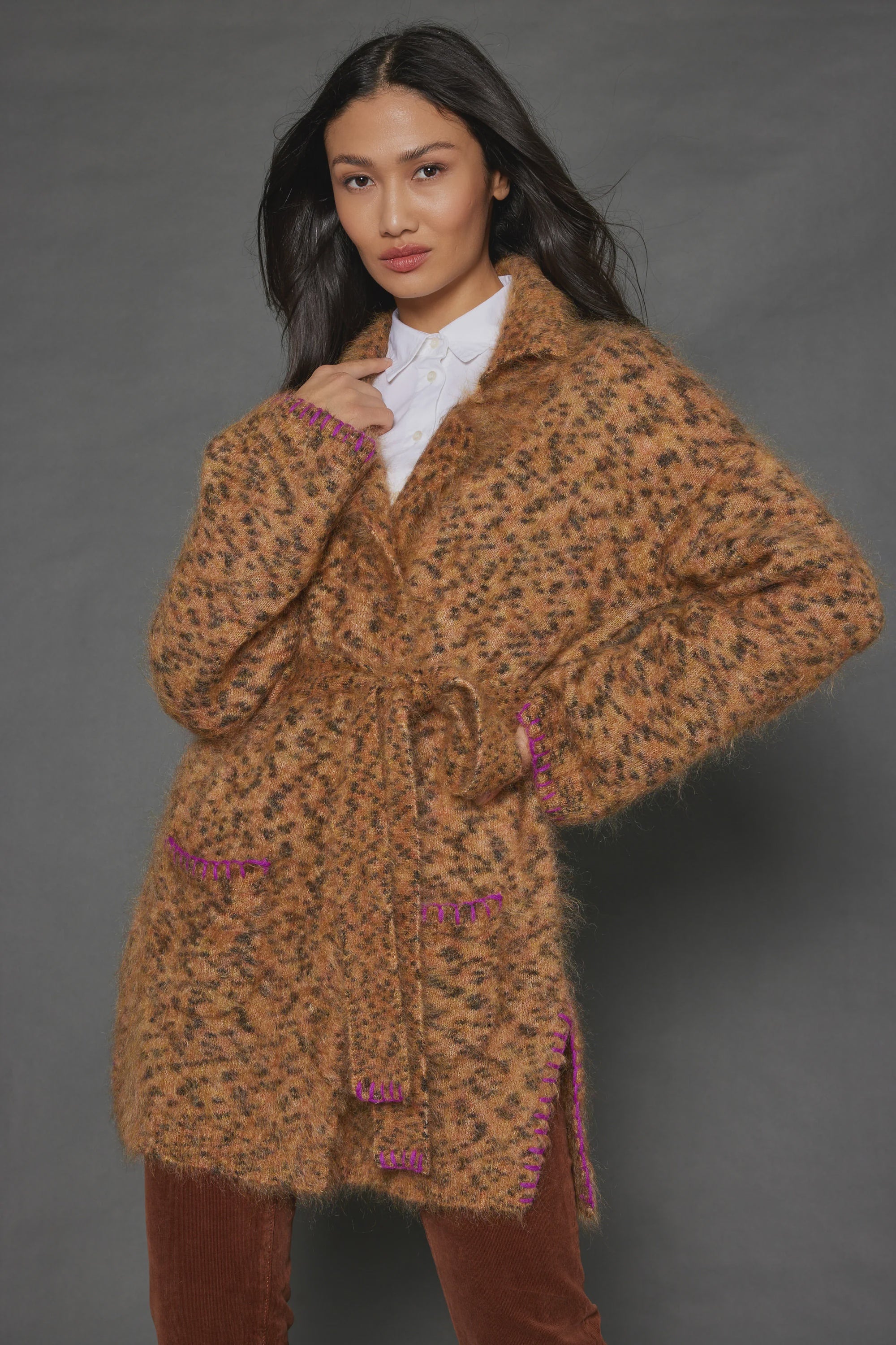 A person poses in the "Mohair Limited Edition" cardigan by Lisa Todd, crafted from luxurious Italian yarn. This textured brown piece is adorned with pink accents at the edges and pockets and includes a tie belt. Beneath it, they wear a white shirt, and their long dark hair cascades over their shoulders against a gray background—showcasing this truly limited edition item.