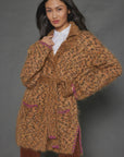 A person poses in the "Mohair Limited Edition" cardigan by Lisa Todd, crafted from luxurious Italian yarn. This textured brown piece is adorned with pink accents at the edges and pockets and includes a tie belt. Beneath it, they wear a white shirt, and their long dark hair cascades over their shoulders against a gray background—showcasing this truly limited edition item.