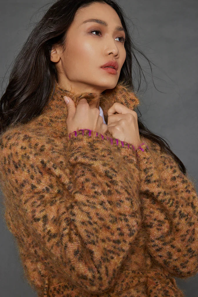 A person with long dark hair holds the collar of the Mohair Limited Edition sweater from Lisa Todd, crafted from exquisite Italian yarn in a fuzzy leopard pattern. The individual gazes upward thoughtfully against a plain, muted gray background.