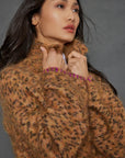 A person with long dark hair holds the collar of the Mohair Limited Edition sweater from Lisa Todd, crafted from exquisite Italian yarn in a fuzzy leopard pattern. The individual gazes upward thoughtfully against a plain, muted gray background.