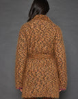 A person with long dark hair is wearing the Lisa Todd Mohair Limited Edition, a luxurious leopard-print coat crafted from fine Italian yarn, over maroon pants. They stand against a plain gray background, facing away from the camera.