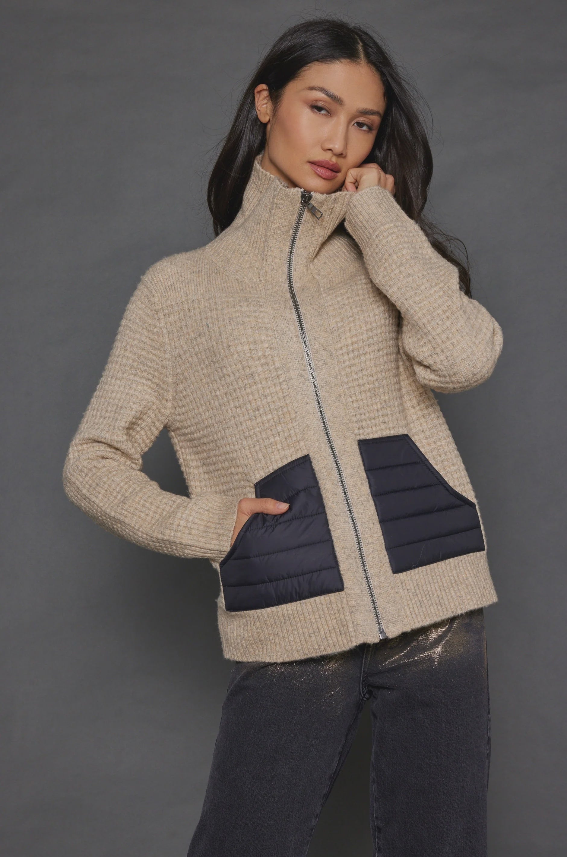 A woman with long dark hair is posing against a gray background, wearing the Deep Pockets Zip by Lisa Todd. This textured stitch cotton blend zip-up sweater features a high collar and two black patch pockets. She pairs it with dark pants and rests one hand on her chin.